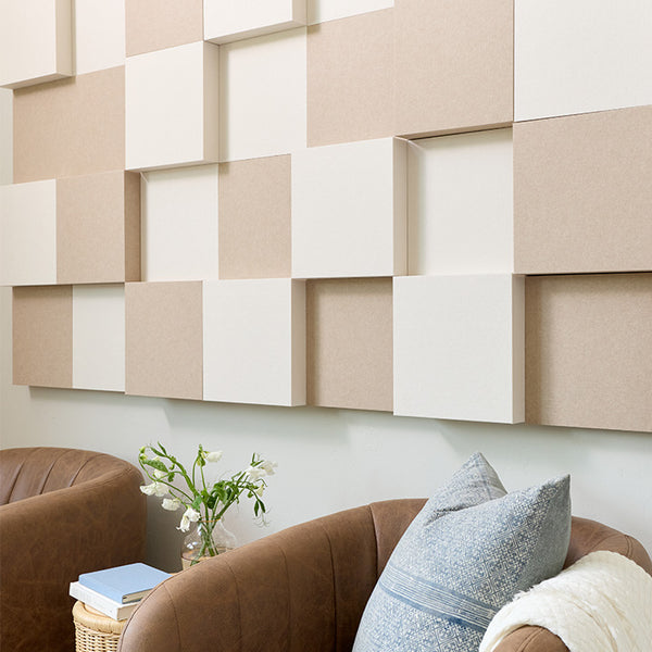 A styled interior scene showing wall-mounted geometric 3D decor in warm beige and off-white tones, installed above brown upholstered chairs with pillows and a small side table with flowers.