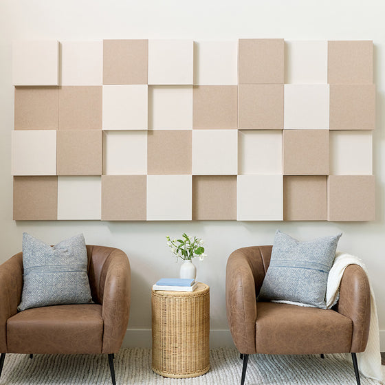 A styled interior scene showing large geometric acoustic wall panels set in beige and off-white mounted above two brown armchairs with blue-gray pillows, a small wicker side table, and a vase of flowers.
