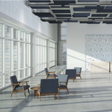 A modern, sunlit office lobby with several blue upholstered wooden armchairs arranged around small round tables. Large floor-to-ceiling windows line the left side, casting strong shadows across the tiled floor.