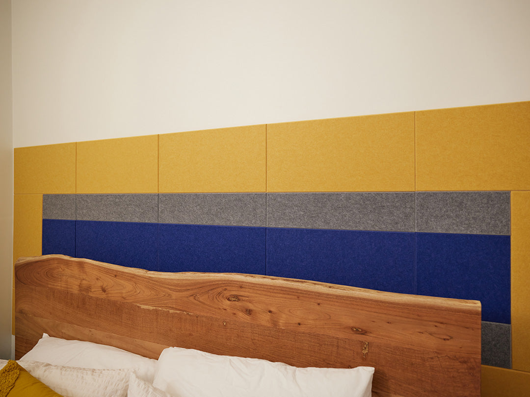 A Felt Right wall design as a headboard in blue yellow and grey felt tiles on a white wall in a bedroom behind a bed 