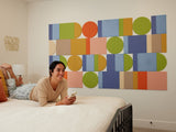 A Felt Right wall art design in shades of blue, green, and orange creating a geometric shape on a cream wall in a bedroom with a bed and a woman laying on it.