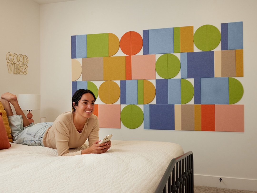A Felt Right wall art design in shades of blue, green, and orange creating a geometric shape on a cream wall in a bedroom with a bed and a woman laying on it.