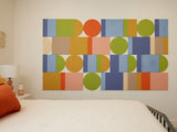 A Felt Right wall art design using geometric shapes in shades of blue, green, yellow, and orange on a cream wall in a bedroom next to a bed.