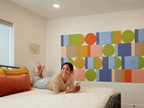 A Felt Right wall art design on a cream wall using geometric shapes in shades of blue, green, and orange along a wall in a bedroom next to a bed with a woman laying on it. 