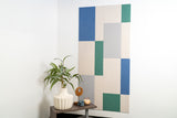 Felt Right wall art design in shades of cream, blues, and green on a white wall next to a little table with a plant and decor.