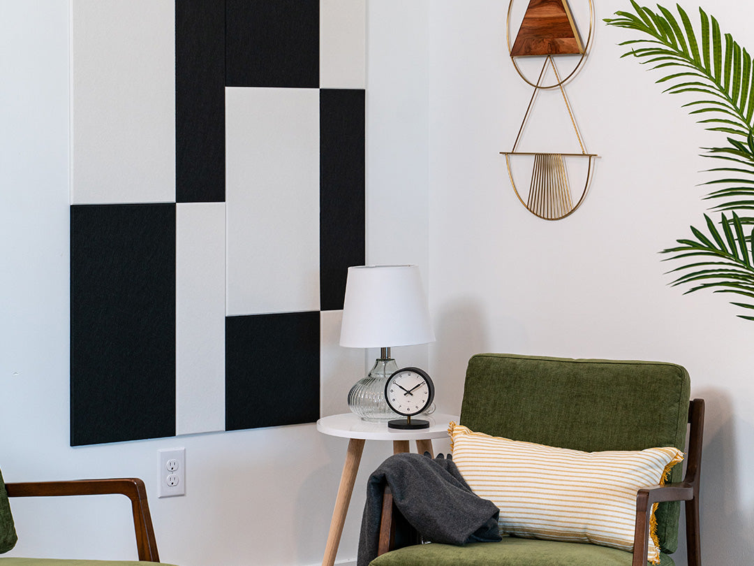 A Felt Right wall art design in black and white rectangles and squares on a white wall next to a side table with a little lamp and clock and two chairs