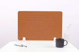 Tan cork desk divider with curved line pattern and white metal support stands on white surface.