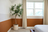 Bedroom wall with white upper paneling and orange-brown lower wainscoting, potted plant, window with curtains, and partial be