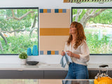 A Felt Right wall art design in shades of yellow, cream, and blue felt tiles in a kitchen in between two windows on a white wall above a counter top.