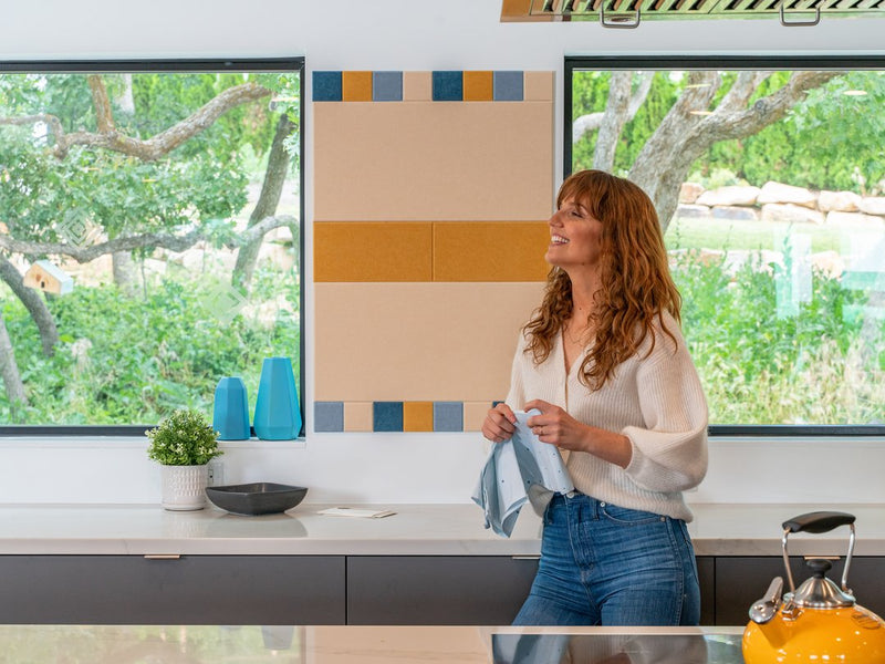 A Felt Right wall art design in shades of yellow, cream, and blue felt tiles in a kitchen in between two windows on a white wall above a counter top.