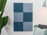 Felt Right wall art design using shades of blue squares with beveled x's on a white wall next to a house plant and a chair.