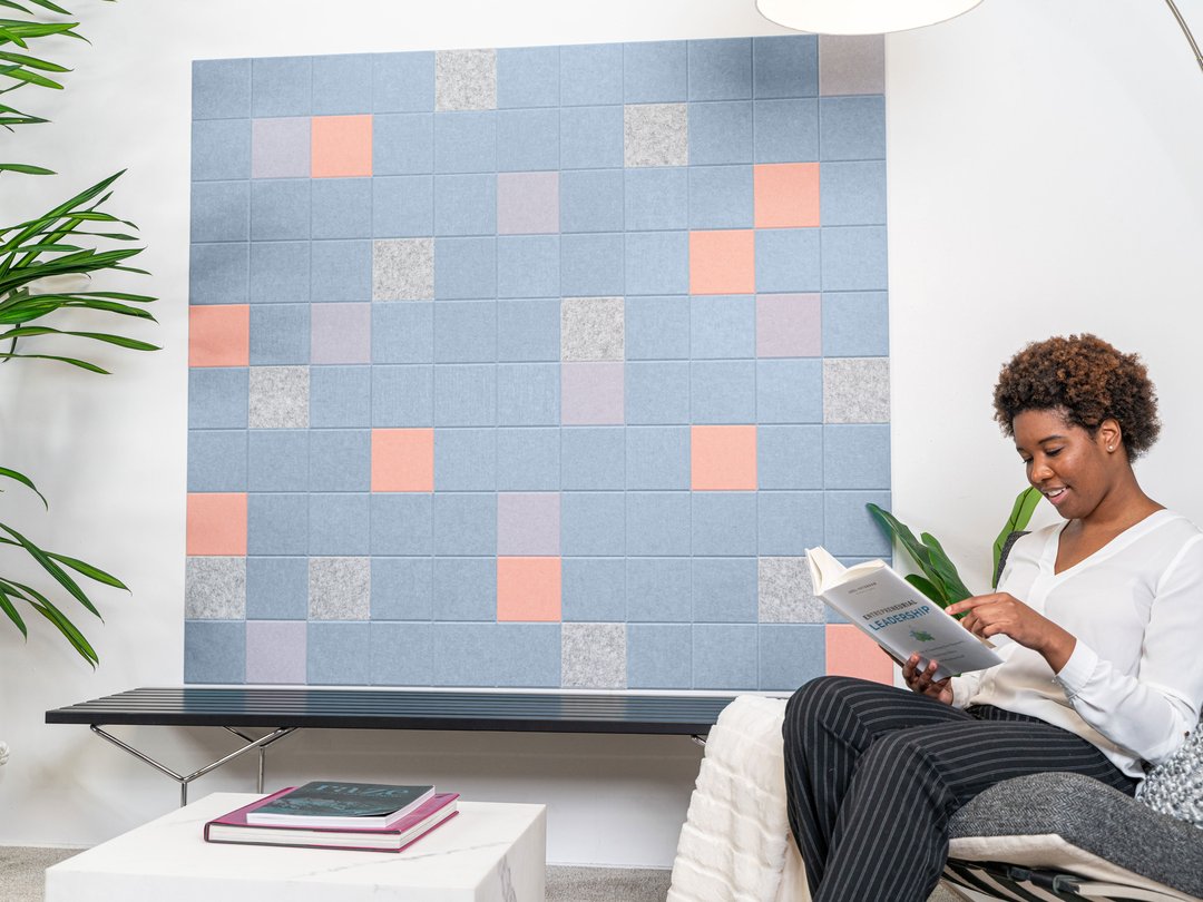 Felt Right wall art design in shades of blue, grey, pink, and purple felt sqaures creating a geometric pattern on a white wall with a plant and a woman reading in the corner.