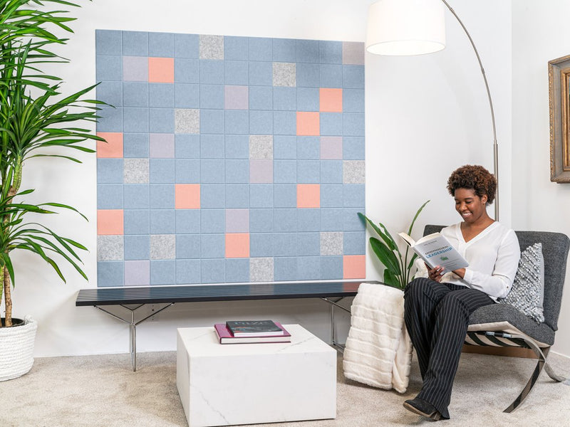 Felt Right wall art design in shades of blue, grey, pink, and purple felt sqaures creating a geometric pattern on a white wall with a plant and a woman reading in the corner.