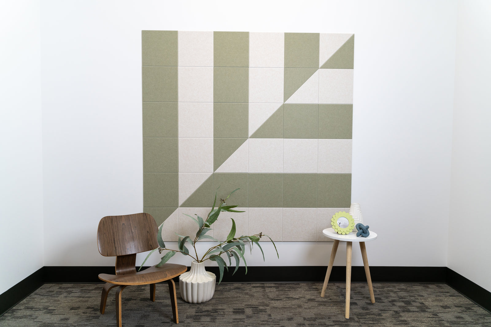 A Felt Right wall art design in shades of cream and green on a white wall behind a chair, plant, and side table.