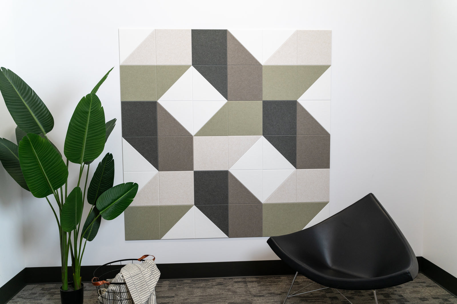 A Felt Right wall art design using shades of green, grey, and cream square and triangle tiles on a white wall next to a chair and a house plant.