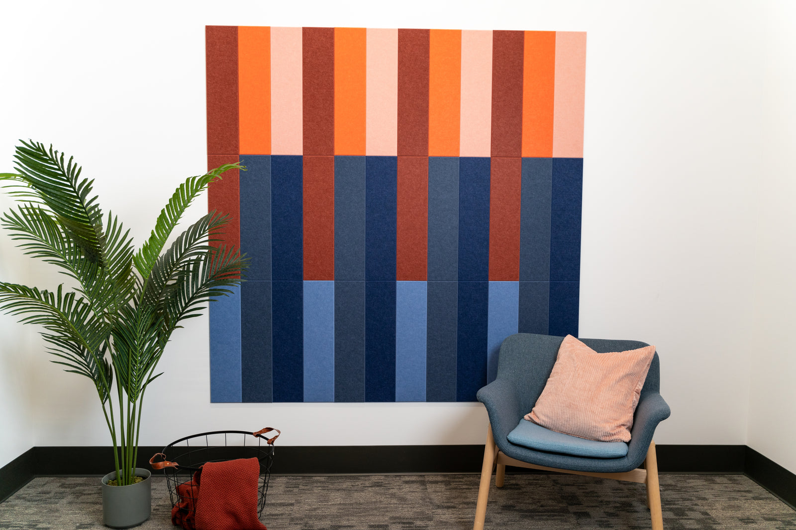 A Felt Right wall art design in shades of blue red and orange on a white wall next to a chair and house plant.