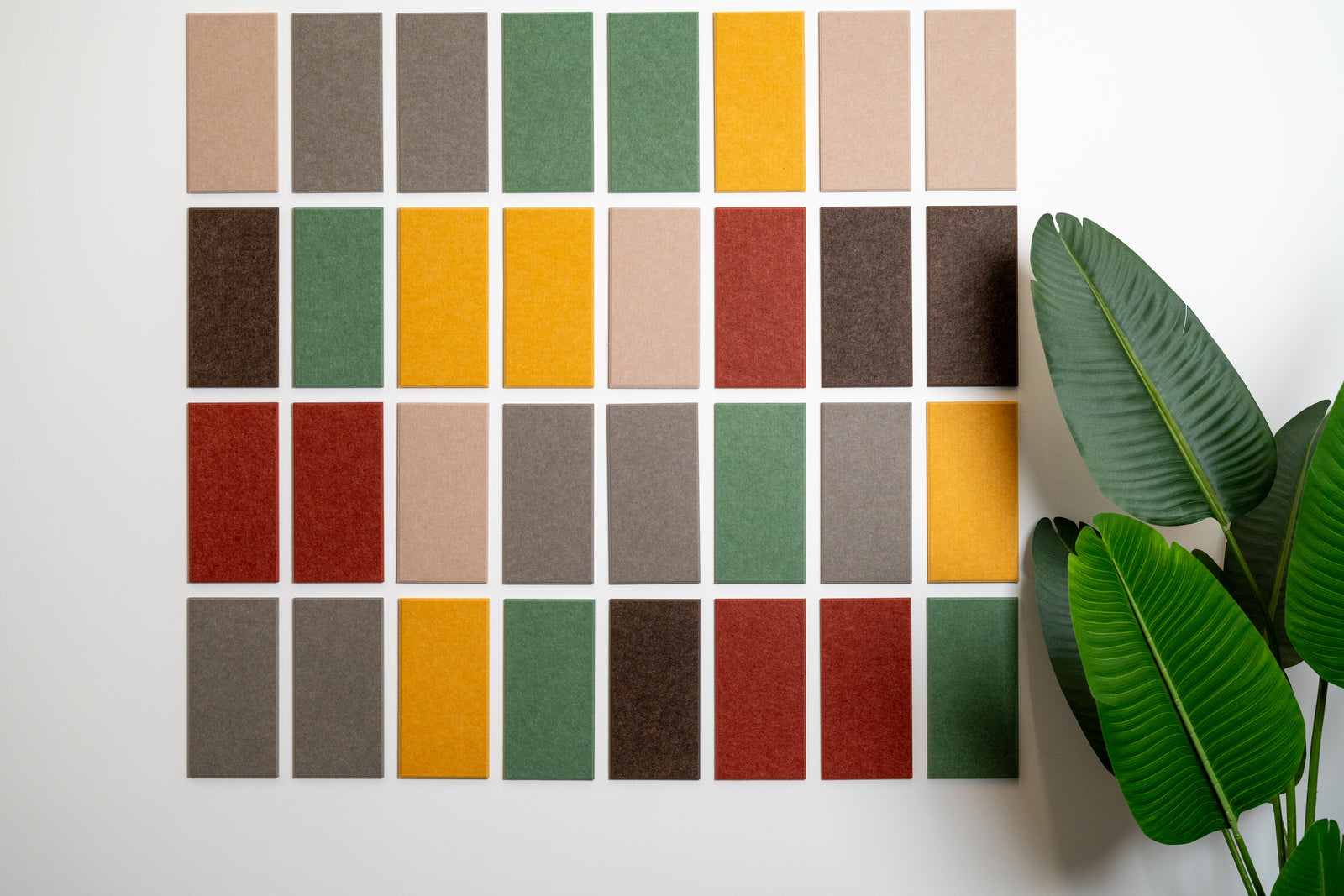A Felt Right wall design in array of solid color sample tiles arranged in a grid pattern on a wall, with a variety of colors including greens, yellows, reds, and grays, and a green plant in the bottom right corner.