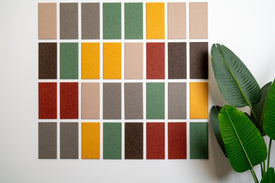 A Felt Right wall design in array of solid color sample tiles arranged in a grid pattern on a wall, with a variety of colors including greens, yellows, reds, and grays, and a green plant in the bottom right corner.