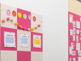 Pink and cream pinboard wall display with circular letter tiles, colorful pom-pom garland, and motivational cards.