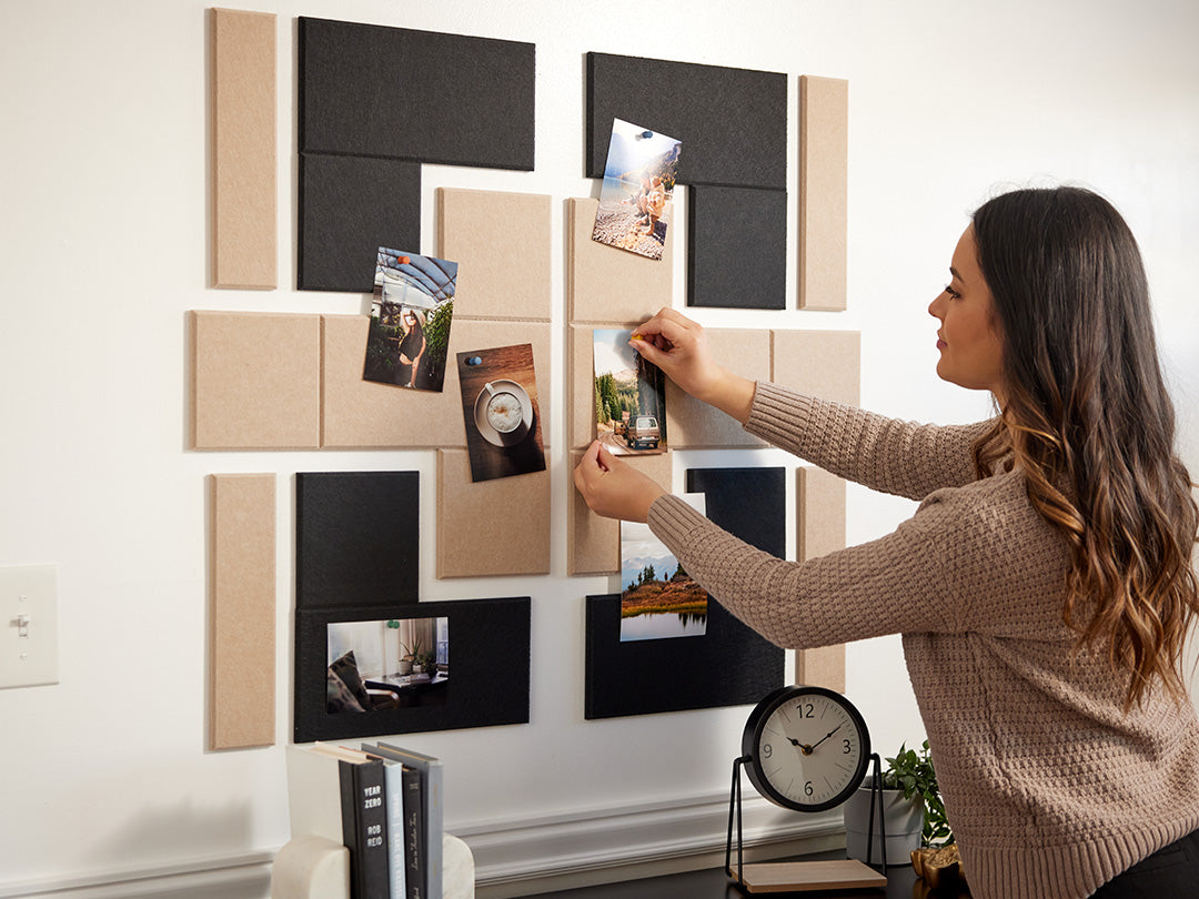 A Felt Right design on a wall above a table with books and a clock on it in shades of tan and black felt tiles that are pinnable with a woman pinning photos on the tiles