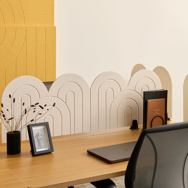 A modern office desk setup featuring a light wood desk, a black mesh office chair, a laptop, a small black vase with dried stems, a black picture frame, and a brown notebook on a desk organizer. The background showcases decorative beige arch acoustic desk dividers against a split cream and mustard yellow wall.