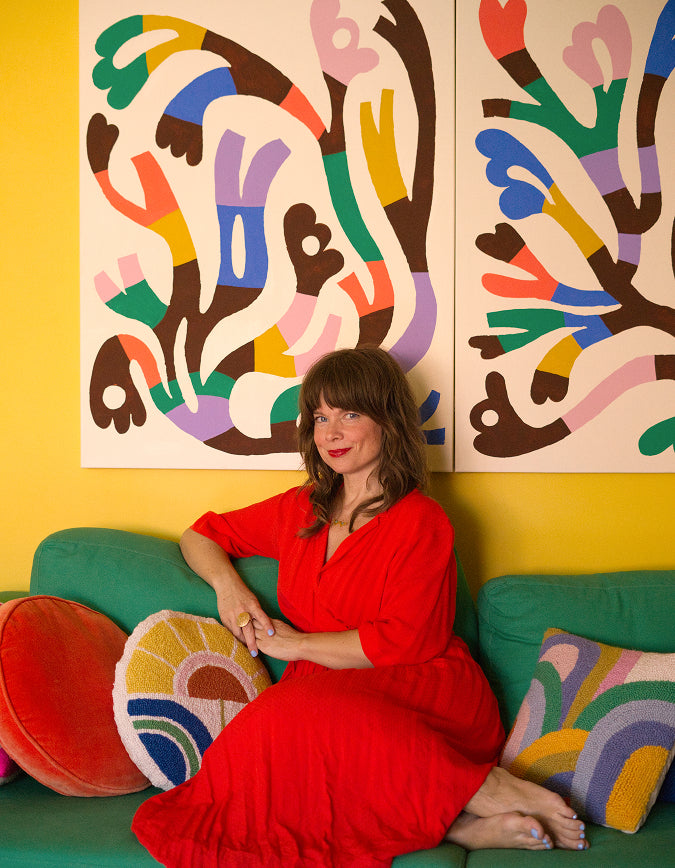A woman in a bright red dress sits on a green sofa in a colorful room, with abstract multicolor wall art hanging above her. She is surrounded by decorative pillows, including a red round pillow and a striped pillow with a rainbow pattern.