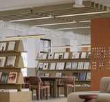 A modern interior space with wooden shelving displaying books, round wooden chairs, linear pendant lights, slatted ceiling panels, and a terracotta-colored partition wall with circular cutouts.