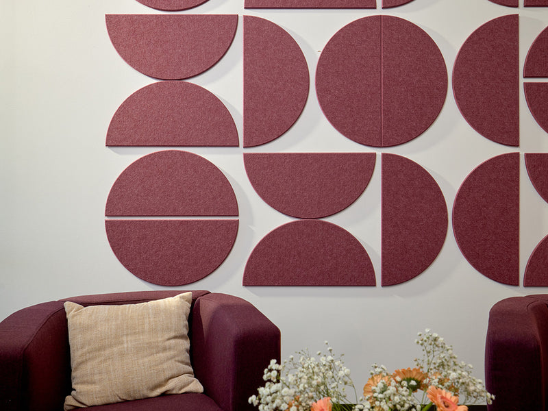 A Felt Right wall art design using largge half circles in a dark red shade on a white wall in an office above a chair with an accent pillow.