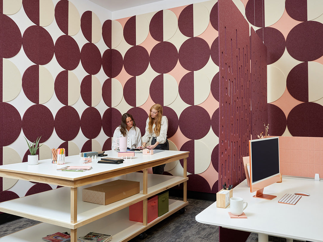 A Felt Right wall art design in shades of pink purple and cream circle tiles creating geometric pattern on a wall corner with a craft table and two women working.