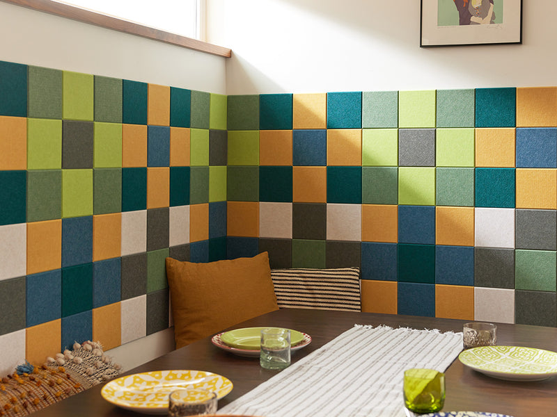A Felt Right wall art design using shades of blue, green, yellow, and grey square felt tiles making a gingham patter along a wall by a kitchen table with decorative pillows