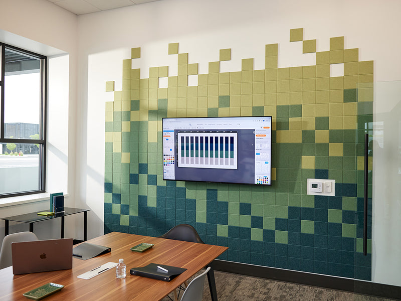 Felt right wall art pixel pattern in shades of green on a white wall in a conference room with a mounted tv on the wall and a big conference table and a window in the corner.