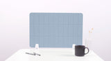 Blue grid desk divider with rounded corners positioned upright on white desk surface with pens and mug.
