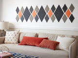 A Felt Right wall art design in grey and orange diamonds on a white wall and a couch with decorative pillows and a lamp on a side table.