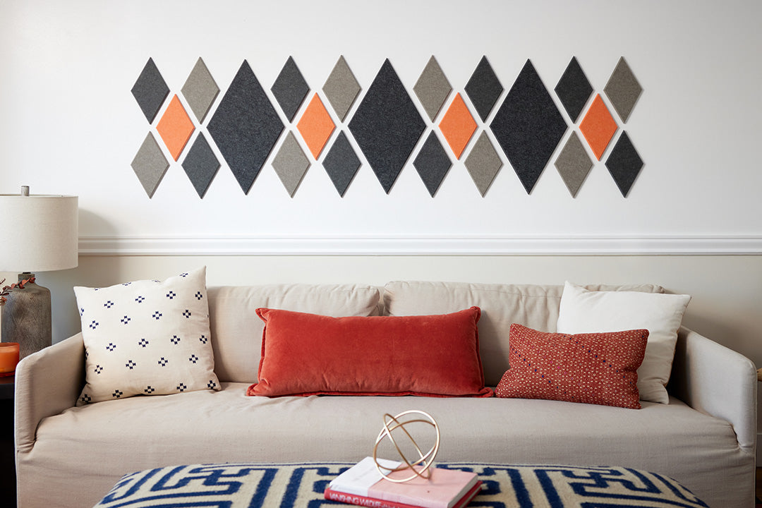 A Felt Right wall art design in grey and orange diamonds on a white wall and a couch with decorative pillows and a lamp on a side table.