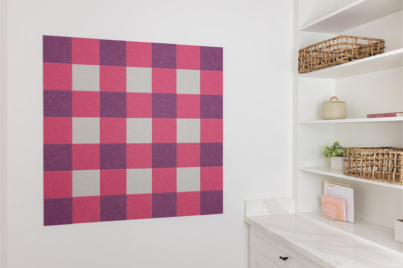 Checkered acoustic wall panel in pink, purple, and gray on a white wall with shelves below.
