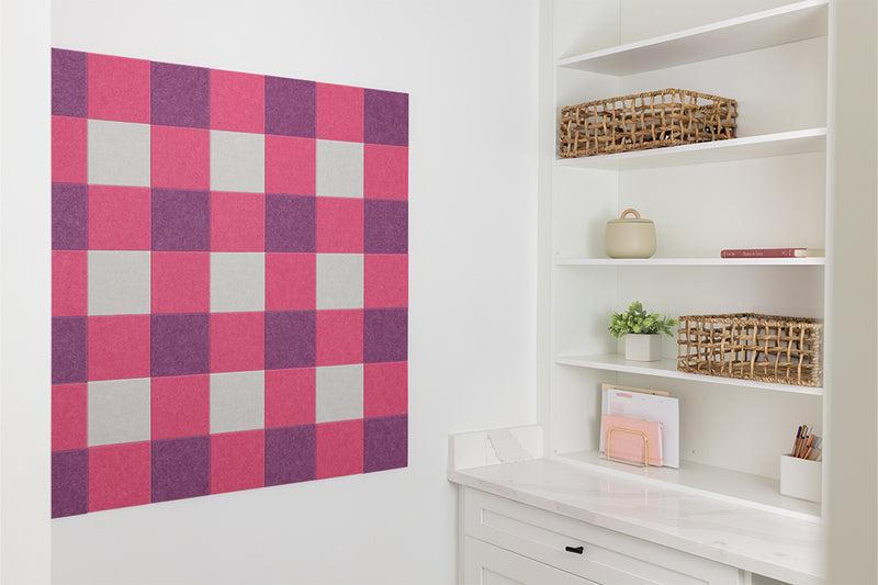 Checkered acoustic wall panel in pink, purple, and gray on a white wall with shelves below.