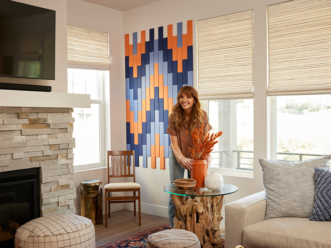 A Felt Right wall art design in shades of blue and orange in felt shiplap like tiles on a cream wall in between some windows in a living room with a fireplace.