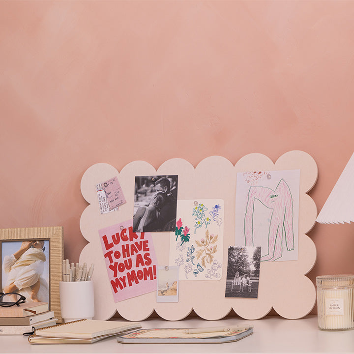 A scalloped-edged light pink display board with clips, styled on a desk against a peach-pink wall. The pinboard is decorated with various notes, photos, and a sketch, alongside lifestyle desk props like a candle, notebook, and picture frame.