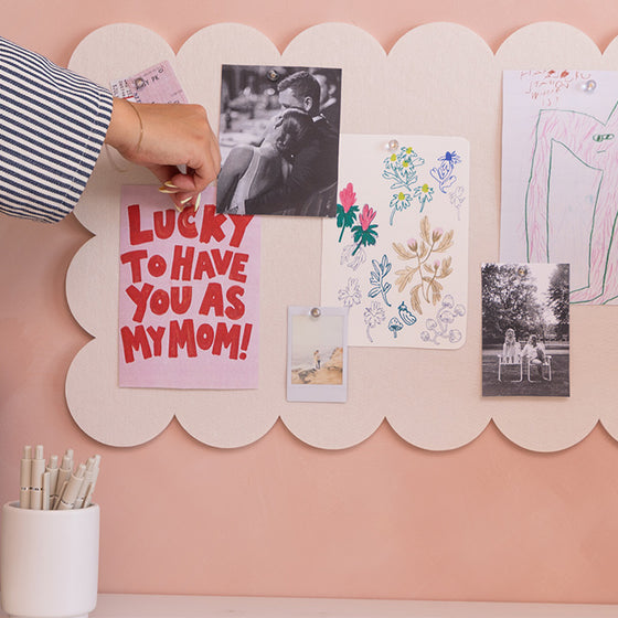A hand with a bracelet is pinning a black-and-white photo onto a large, scalloped-edged cream-colored pinboard mounted on a dusty pink wall. The felt board is decorated with various keepsakes, including a pink card that reads 'LUCKY TO HAVE YOU AS MY MOM!' in red letters, a floral drawing, and a child's colored pencil sketch.
