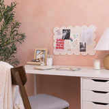A styled home office scene with a white desk against a dusty pink wall. A scalloped felt pinboard on the wall holds several small prints and a card reading 'Lucky to Have You As My Mom!'.