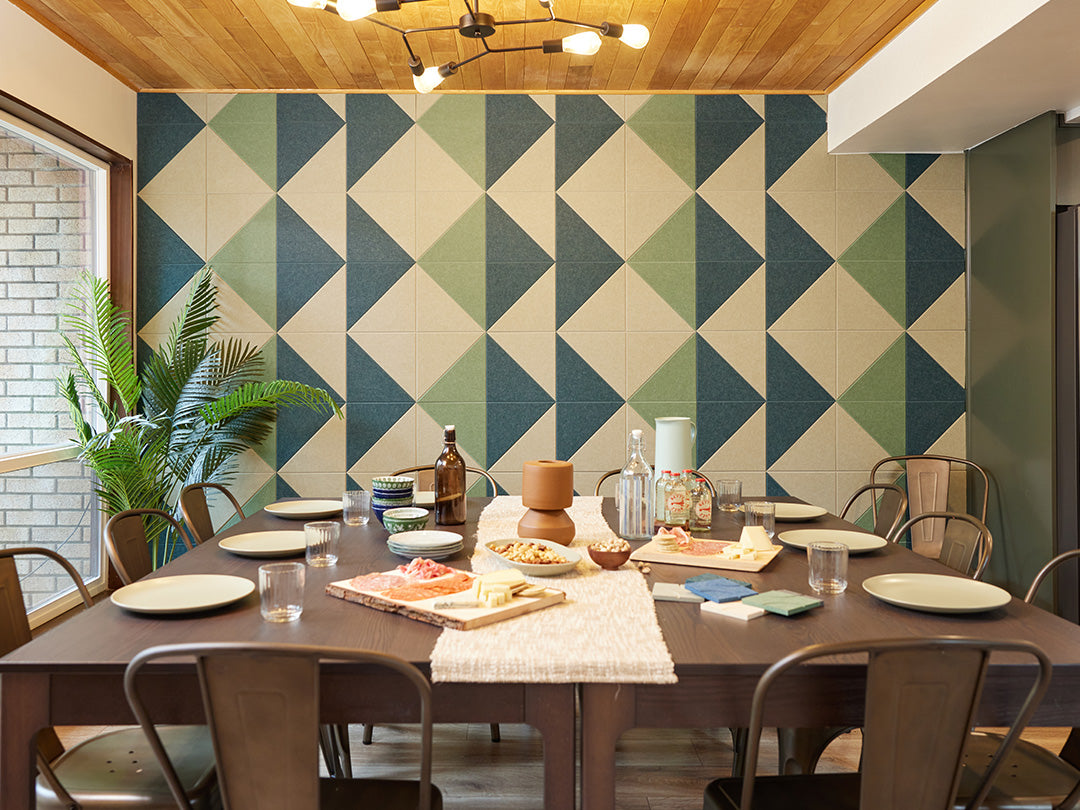 A Felt Right wall art design in shades of green and cream triangles tiles covering a wall in a dinning room with a set dinning table.