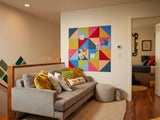 Felt Right wall art design using triangular felt tiles in the shades of pink, red, green, and blue with card pinned to it next to a couch with decorative pillows.
