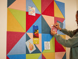 Felt Right wall art design using triangular felt tiles in the shades of pink, red, green, and blue with a woman pinning a card to it.