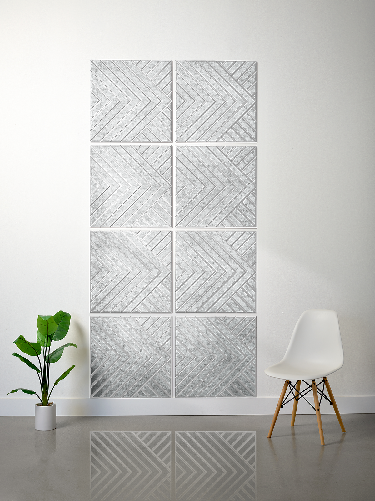 A modern wall art piece made of nine square panels arranged in a 3x3 grid, featuring a textured silver and white chevron pattern. The panels are mounted on a white wall in a minimalist room setting with a small green potted plant on the left and a white molded chair with wooden legs on the right.