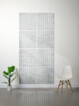 A modern interior scene showing a large wall-mounted grid artwork made of multiple rectangular panels with a light gray and white textured vertical pattern. A white chair with wooden legs sits to the right, and a green potted plant is to the left, against a white wall and gray floor.
