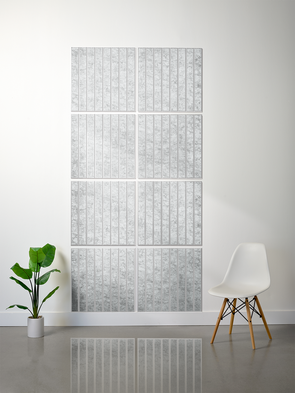 A modern interior scene showing a large wall-mounted grid artwork made of multiple rectangular panels with a light gray and white textured vertical pattern. A white chair with wooden legs sits to the right, and a green potted plant is to the left, against a white wall and gray floor.