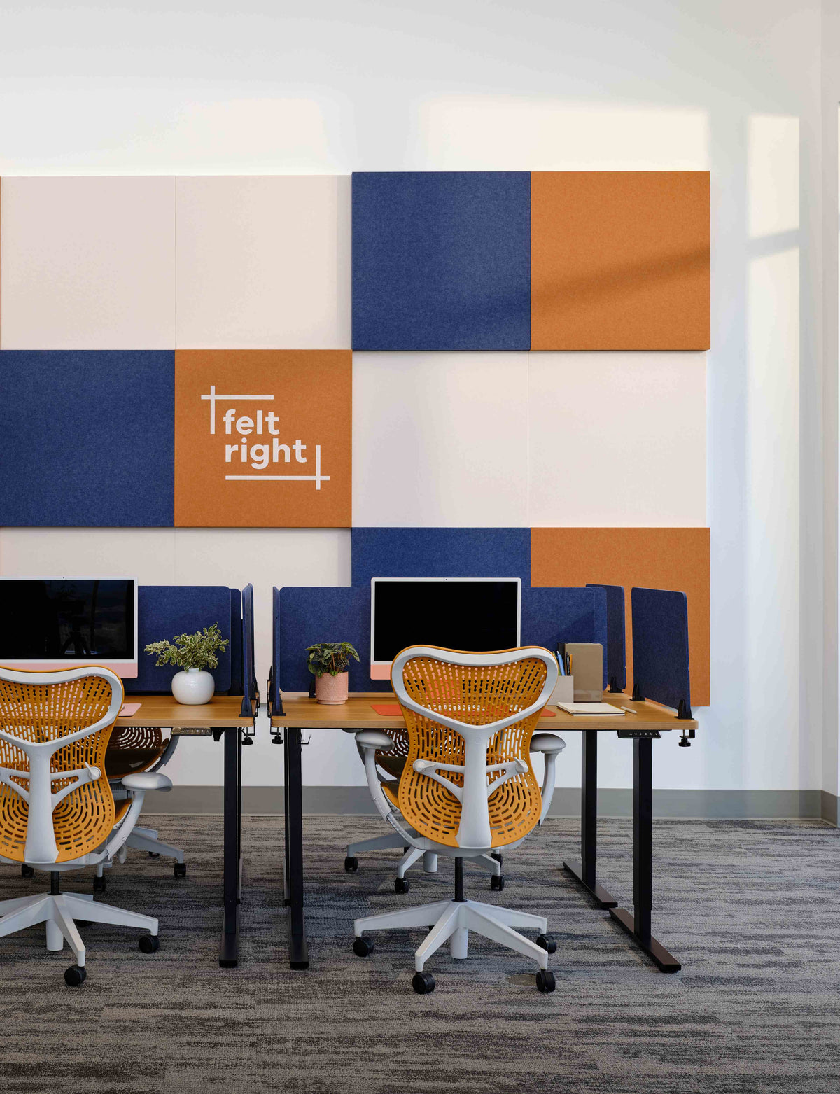 A modern office interior with rows of desks, computer monitors, orange and blue mesh office chairs, and a large geometric wall panel set in cream, navy, and orange. The center panel shows the white text 'felt right'.