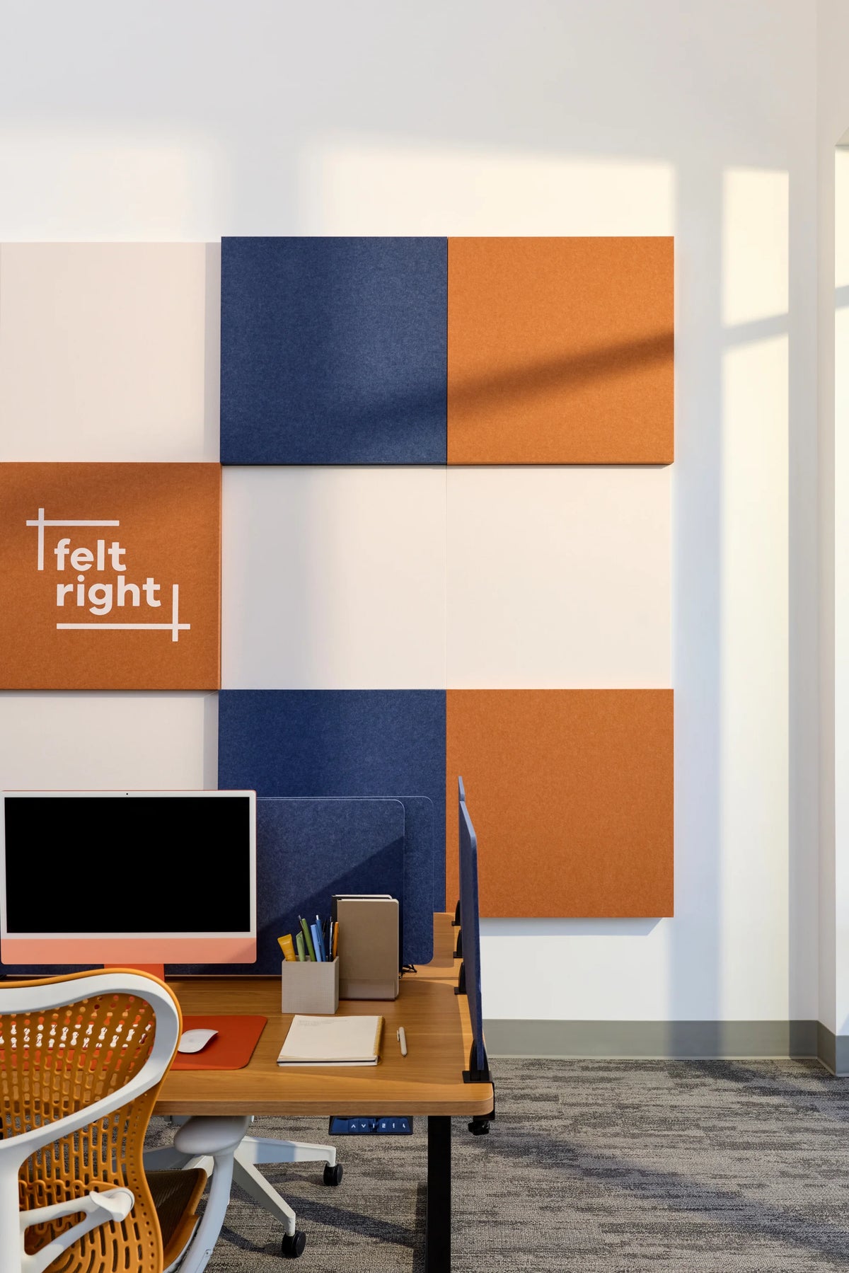 A modern office interior showing a set of felt acoustic wall panels in orange and navy blue arranged in a grid on a white wall. A wooden desk with a computer monitor, office chair, and desk accessories are visible in the foreground.