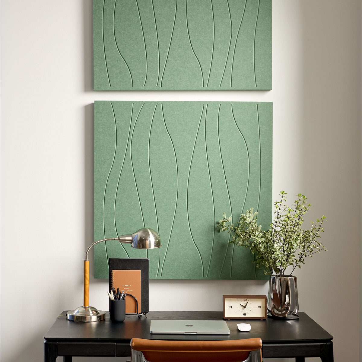 A large rectangular wall art panel with a sage green textured background and thin black wavy line pattern, shown hanging on a light wall in a styled interior with a desk lamp, notebook, plant, and small clock.