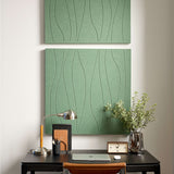 A large rectangular wall art panel with a sage green textured background and thin black wavy line pattern, shown hanging on a light wall in a styled interior with a desk lamp, notebook, plant, and small clock.